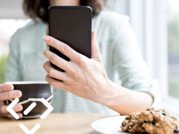 Frau sitzt mit Smartphone in der Hand am Tisch und trinkt Kaffee