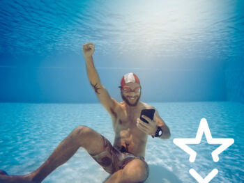 Mann sitz am Grund eines Schwimmbeckens mit dem Smartphone in der Hand und freut sich