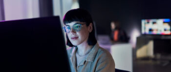 Frau mit dunklen, kurzen Haaren und Brille sitzt im Dunklen vor leuchtendem Bildschirm