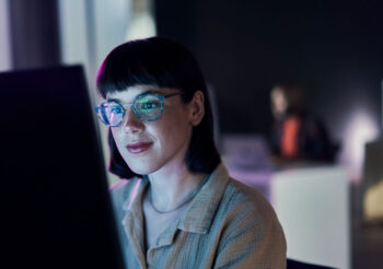 Frau mit dunklen, kurzen Haaren und Brille sitzt im Dunklen vor leuchtendem Bildschirm